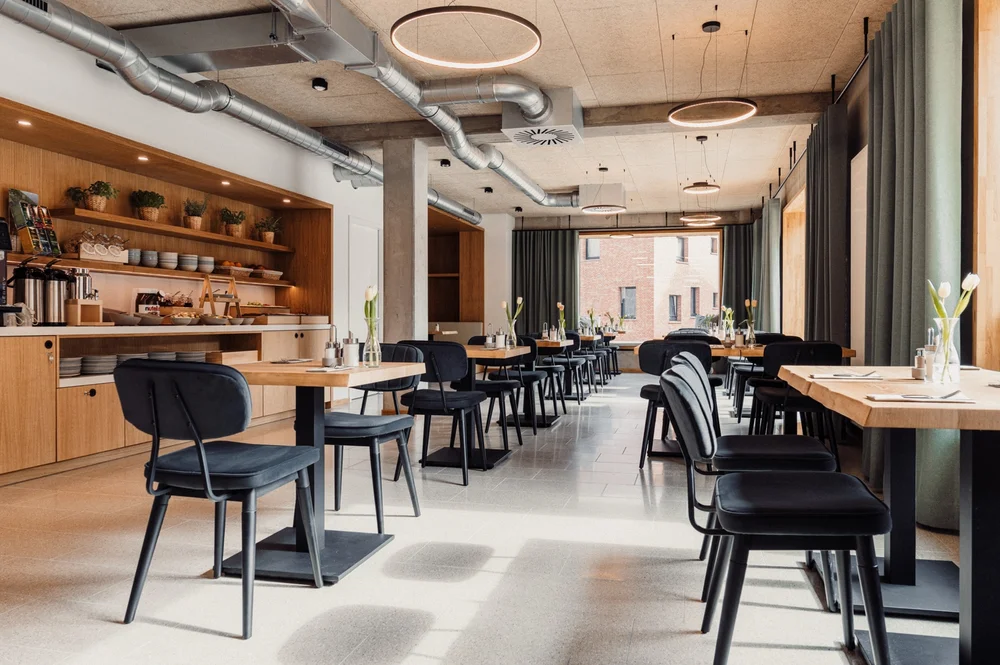 Gemütlicher Frühstücksraum mit gedeckten Holztischen, modernen schwarzen Stühlen, großen Fenstern und Buffetzeile in heller Holzoptik.