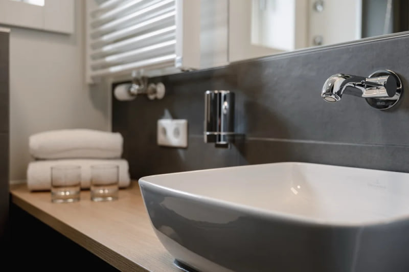 Detailaufnahme eines modernen Badezimmers mit eckigem Waschbecken, verchromtem Wandwasserhahn, Handtüchern und Gläsern auf einer Holzablage.